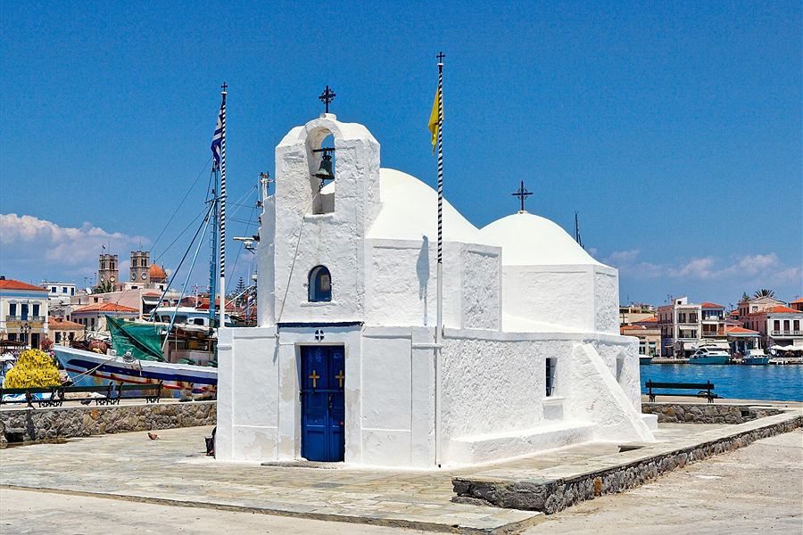 Aegina ferry: a whitewashed harbourside chapel with a blue door, bell gable and two domes stands on a stone quay, with a fishing boat, waterfront houses and the sea behind it.