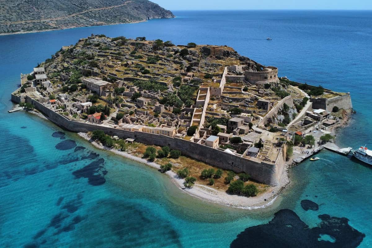 Agios Nikolaos day trip: an aerial view of a whole island fortress with long stone walls, a round bastion and rows of ruined stone houses on a rocky hill, surrounded by turquoise sea, with a white boat at a small quay on the right and a mountain coast behind.