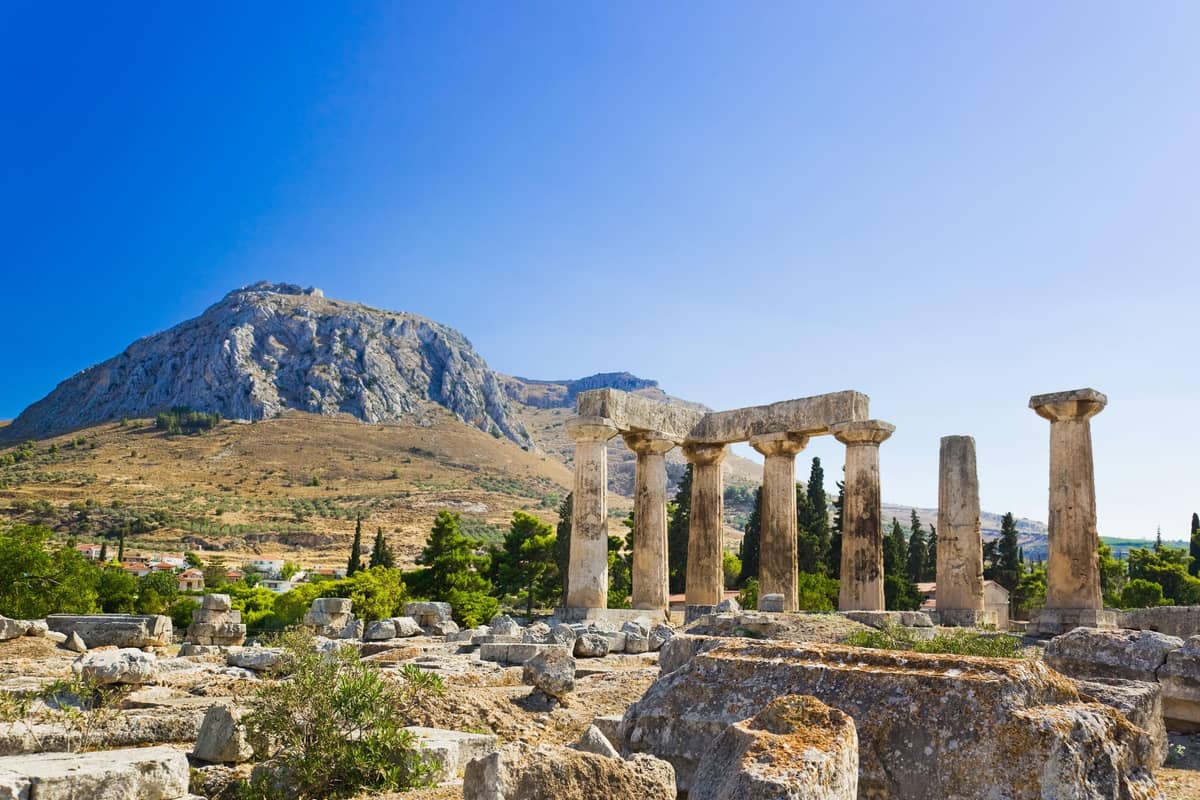 Ancient Corinth day trip: a row of weathered Doric columns with a stone beam on top stands among scattered ruins, with a bare rocky mountain rising behind it and a clear deep blue sky above.