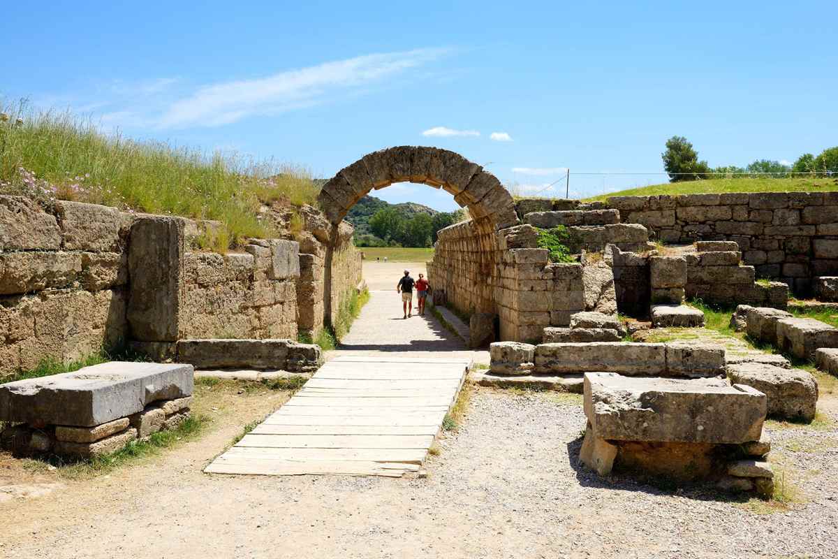 Ancient Olympia tour: two visitors walk through the stone arch of the stadium entrance tunnel