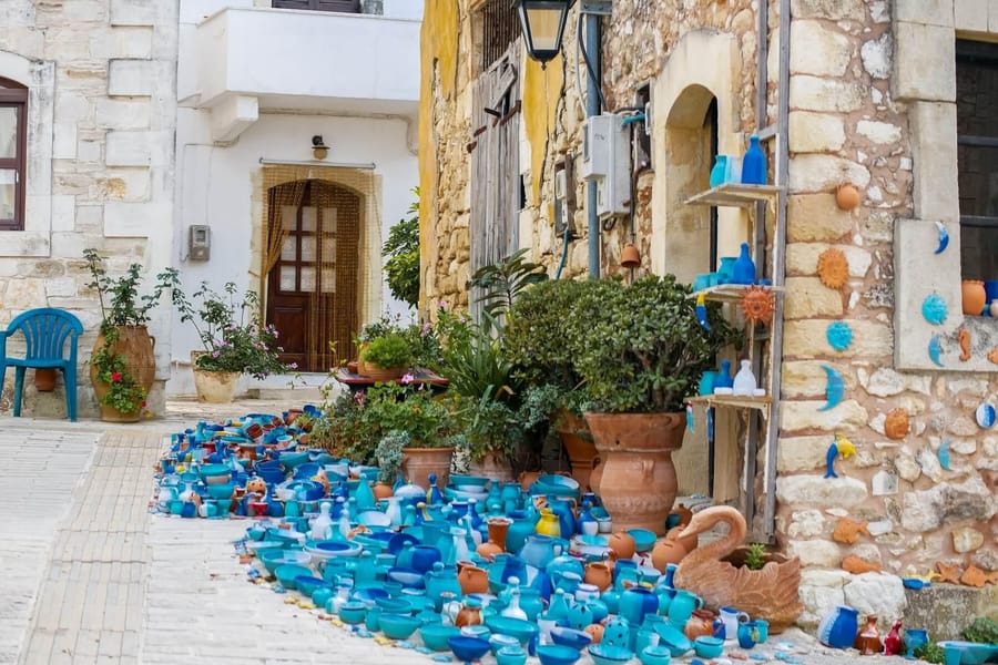 Arkadi monastery tour: blue and turquoise pottery bowls and jugs cover a paved village lane beside a stone house with terracotta pots, green plants and a wooden shelf of ceramic ornaments.