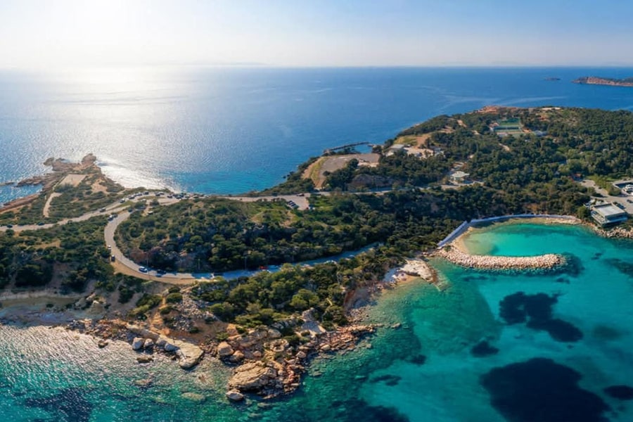 Athens beach trip: an aerial view of a pine-covered headland with a winding road, a turquoise bay with a stone breakwater and a small sandy cove, and sparkling blue sea reaching the horizon.