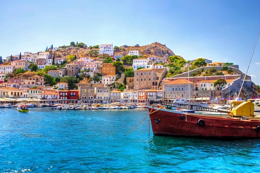 Athens boat trip: a hillside harbour town of stone mansions and white and terracotta-roofed houses rises above bright turquoise water, with small boats moored along the quay and a red wooden boat in the foreground under a clear blue sky.