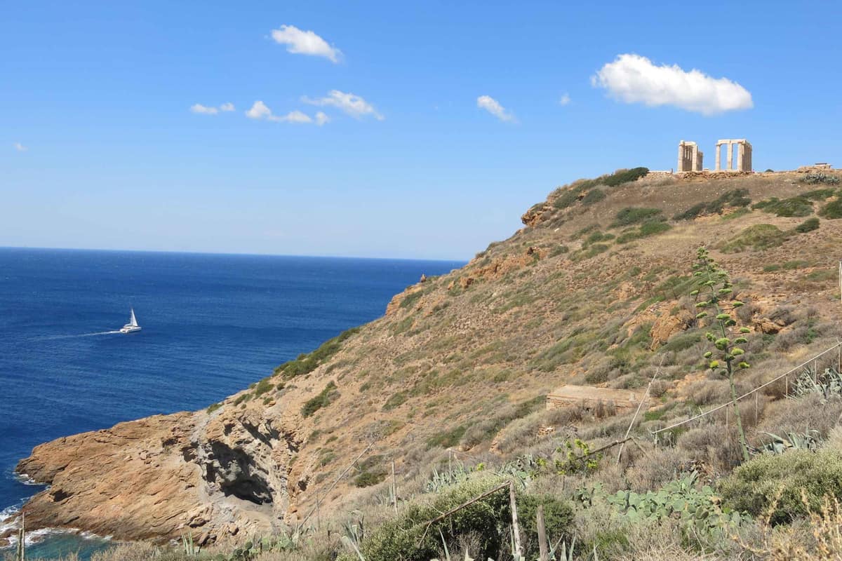 Athens boat trip: a white sailing catamaran leaves a long wake on deep blue sea below a rocky headland, with the columns of the Temple of Poseidon standing on the cape under a bright blue sky with a few small clouds.
