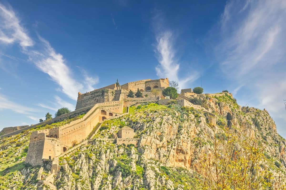 Athens castle tour: a long stone fortress with stepped walls and bastions crowns a steep green and rocky hill under a deep blue sky with wispy white clouds.