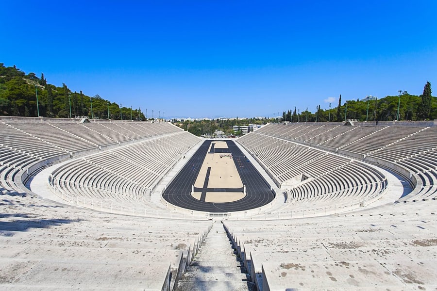 Athens city pass: the white marble seating of the Panathenaic Stadium curves around a dark running track, with pine-covered hills and a blue sky behind.