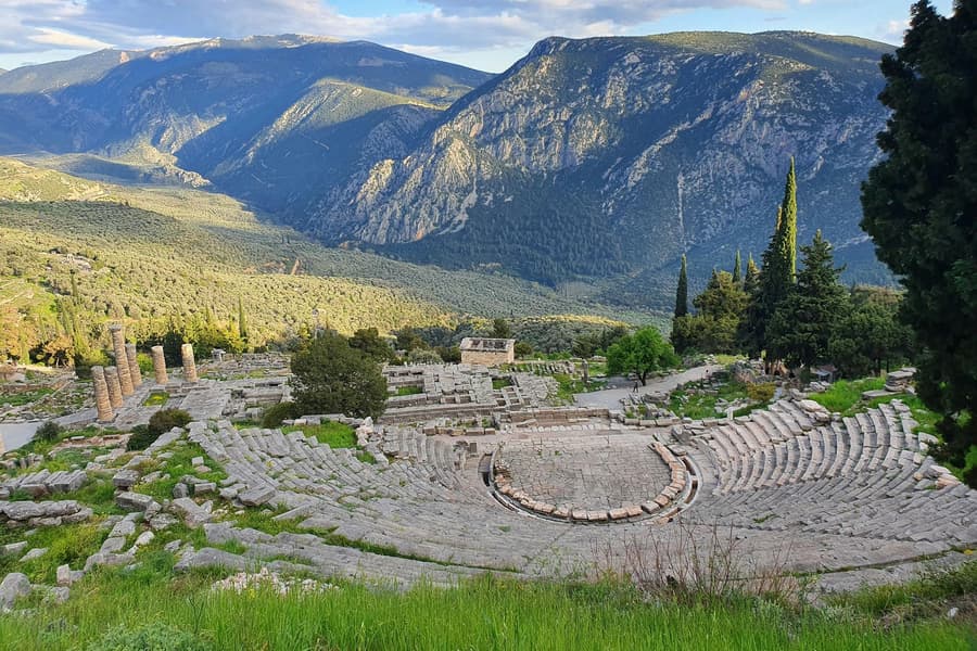 Athens day trip: the curved stone seating of an ancient theatre rests on a green hillside above a wide mountain valley, with tall cypress trees on the right and a rocky ridge behind.