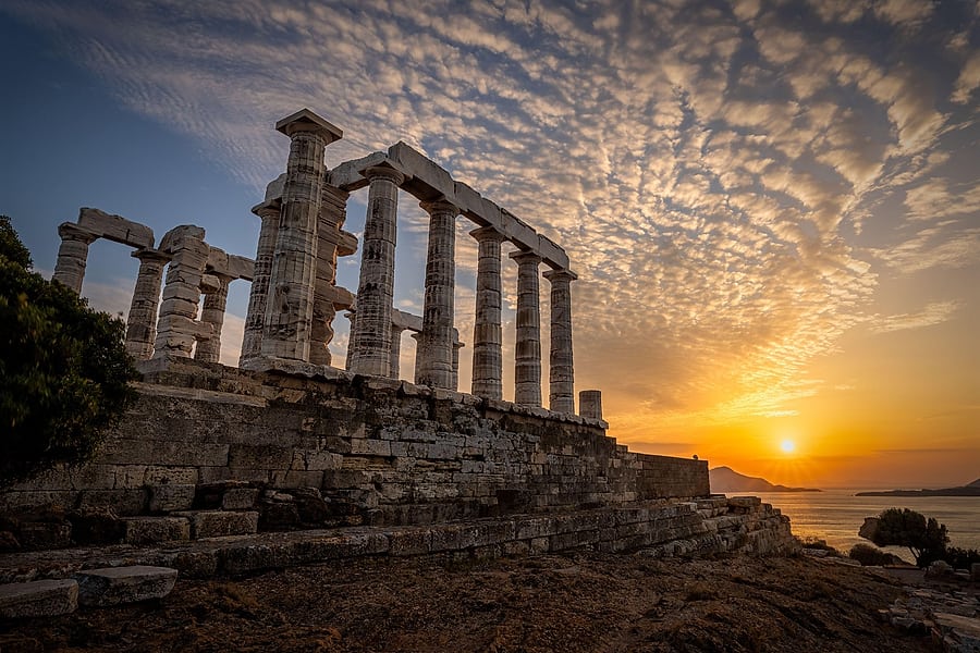 Athens sunset cruise: the white marble columns of a ruined Doric temple stand on a stone terrace under a sky of rippled clouds, with the low orange sun above a calm sea and distant headlands on the right.