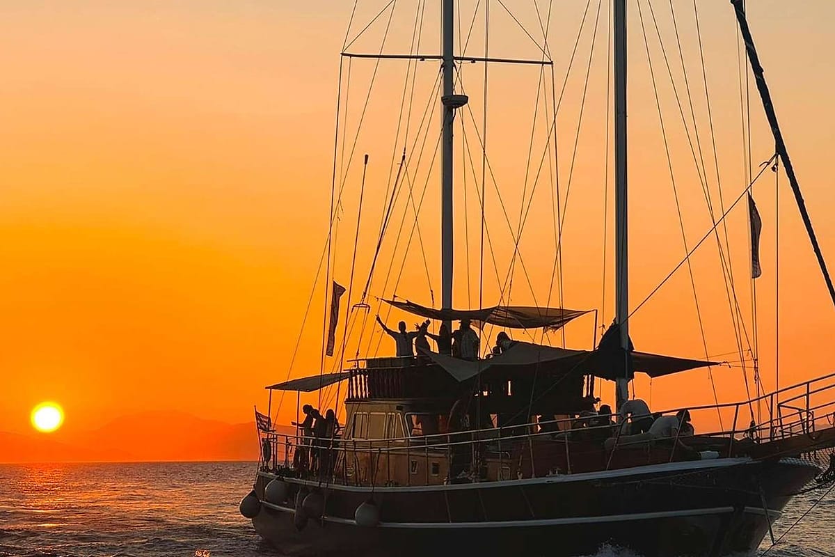 Athens sunset cruise: a dark wooden two-masted sailing ship with its rigging in silhouette against a glowing orange sky, small figures on deck and the low sun above the sea beside hazy hills on the horizon.