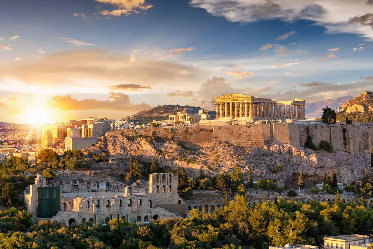 Athens tickets: the columned Parthenon crowns the rocky Acropolis hill above a ruined stone theatre with rows of arched openings and green trees, under a blue sky with golden clouds and a low setting sun.