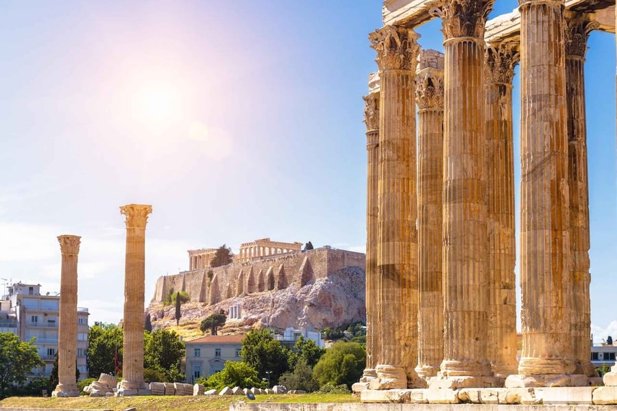 Athens tickets: tall fluted stone columns with carved leaf capitals rise on the right in bright sun, two single columns stand on the left, and the rocky Acropolis with a columned temple on top rises behind green trees under a blue sky with a soft sun glare.
