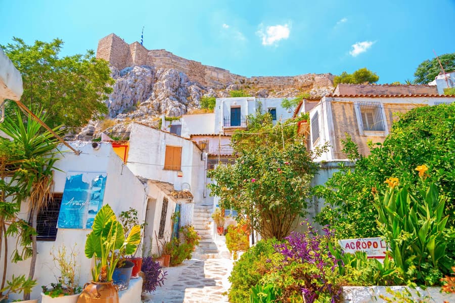 Athens walking tour: a whitewashed stepped lane winds between white houses, potted plants and flowering bushes, with a small signpost on the right, below a rocky hill topped by a stone fortress wall under a bright blue sky.