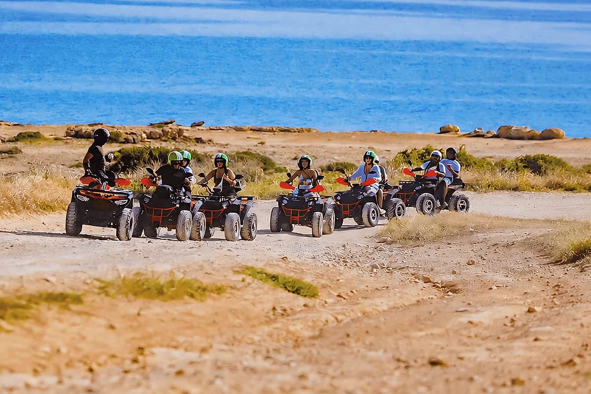 Ayia Napa quad safari: a line of quad bikes with helmeted riders on a pale dirt track edged with dry golden grass, with the deep blue sea stretching behind them under a bright sky.