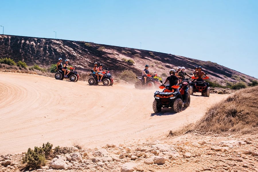 Ayia Napa quad safari: helmeted riders on quad bikes ride in a line round a wide curve of a pale sandy track below a dry hillside, with dust in the air under a clear blue sky.