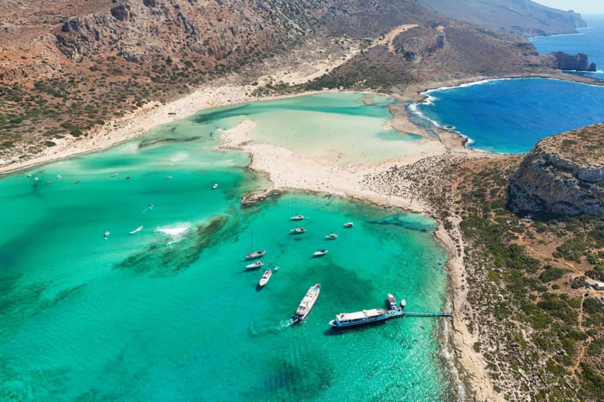 Balos lagoon cruise ship moored at the jetty in Balos bay, seen from above, with yachts at anchor, turquoise shallows and white sand beyond