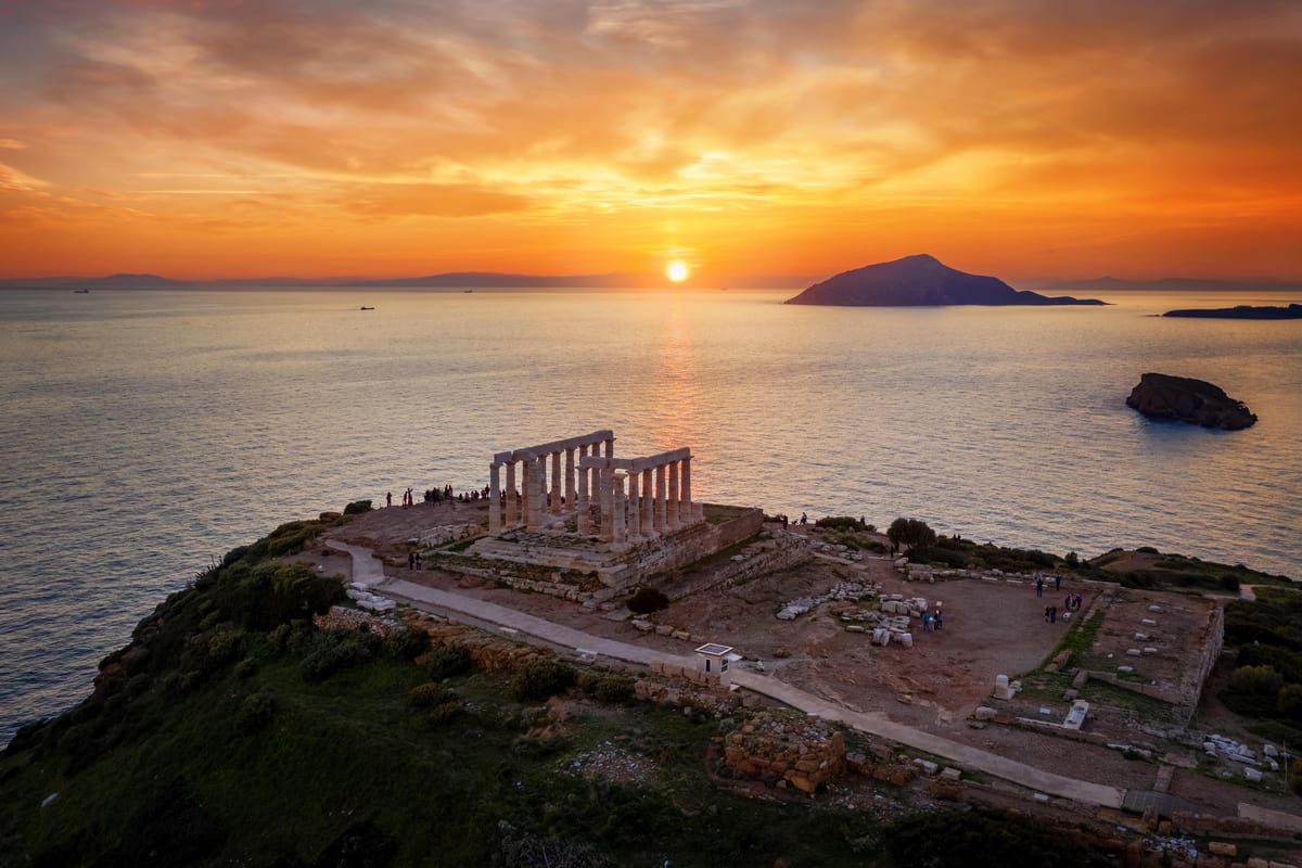 Cape Sounion castle tour: the Doric columns of a white temple on a cliff-top headland at sunset, with the sun on the horizon, calm sea on both sides and a dark island beyond.