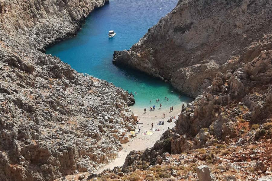 Chania beach trip: looking down between tall grey limestone walls at a narrow cove where a small pebble beach with a few swimmers and towels meets a pocket of turquoise water with a white motorboat.