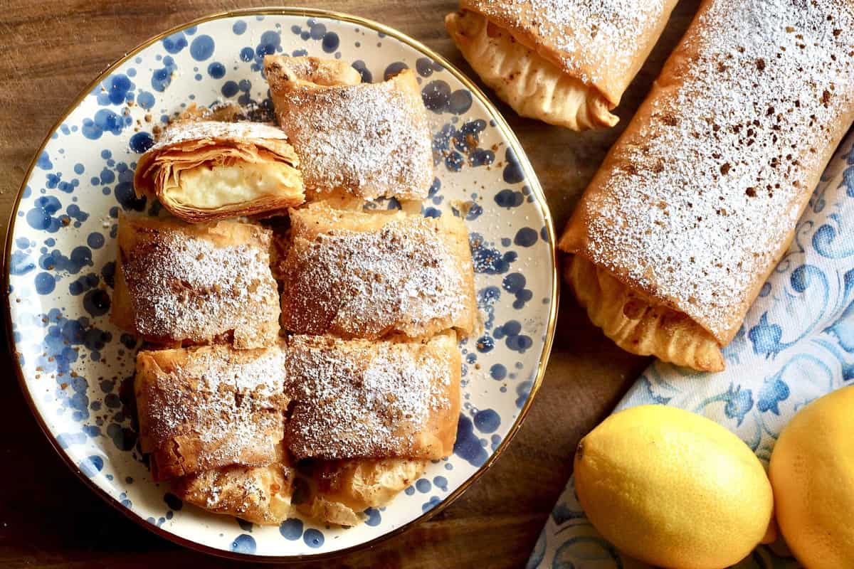 Chania cooking class: golden filo pastry rolls dusted with icing sugar sit on a blue-and-white speckled plate, one piece cut open to show a creamy filling, with two more whole rolls on a wooden table beside a blue patterned cloth and two lemons.