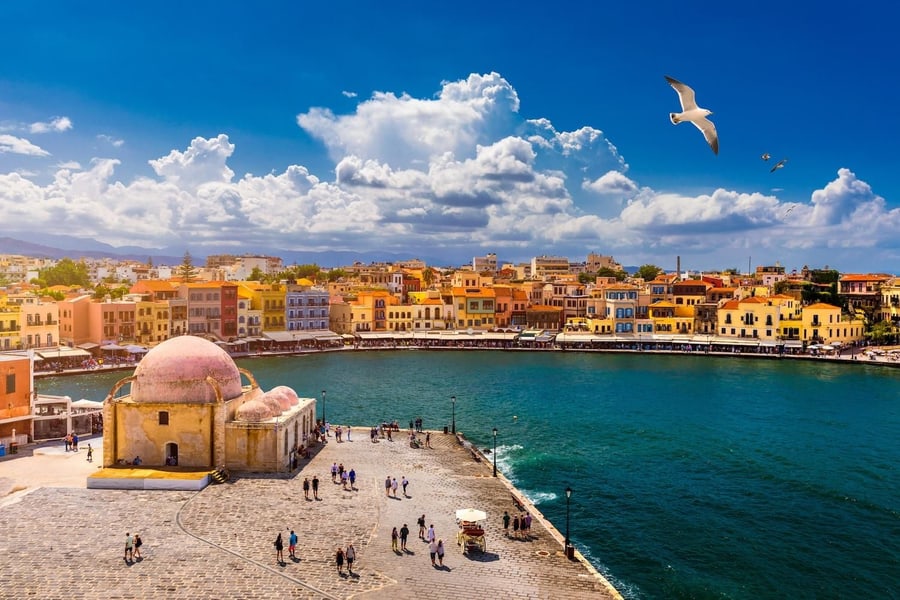 Chania cooking class: a sandy-stone building with a pink dome on a paved quay beside a curving harbour of deep green-blue water, with yellow, orange and pink houses along the waterfront, a seagull in the air and white clouds in a deep blue sky.