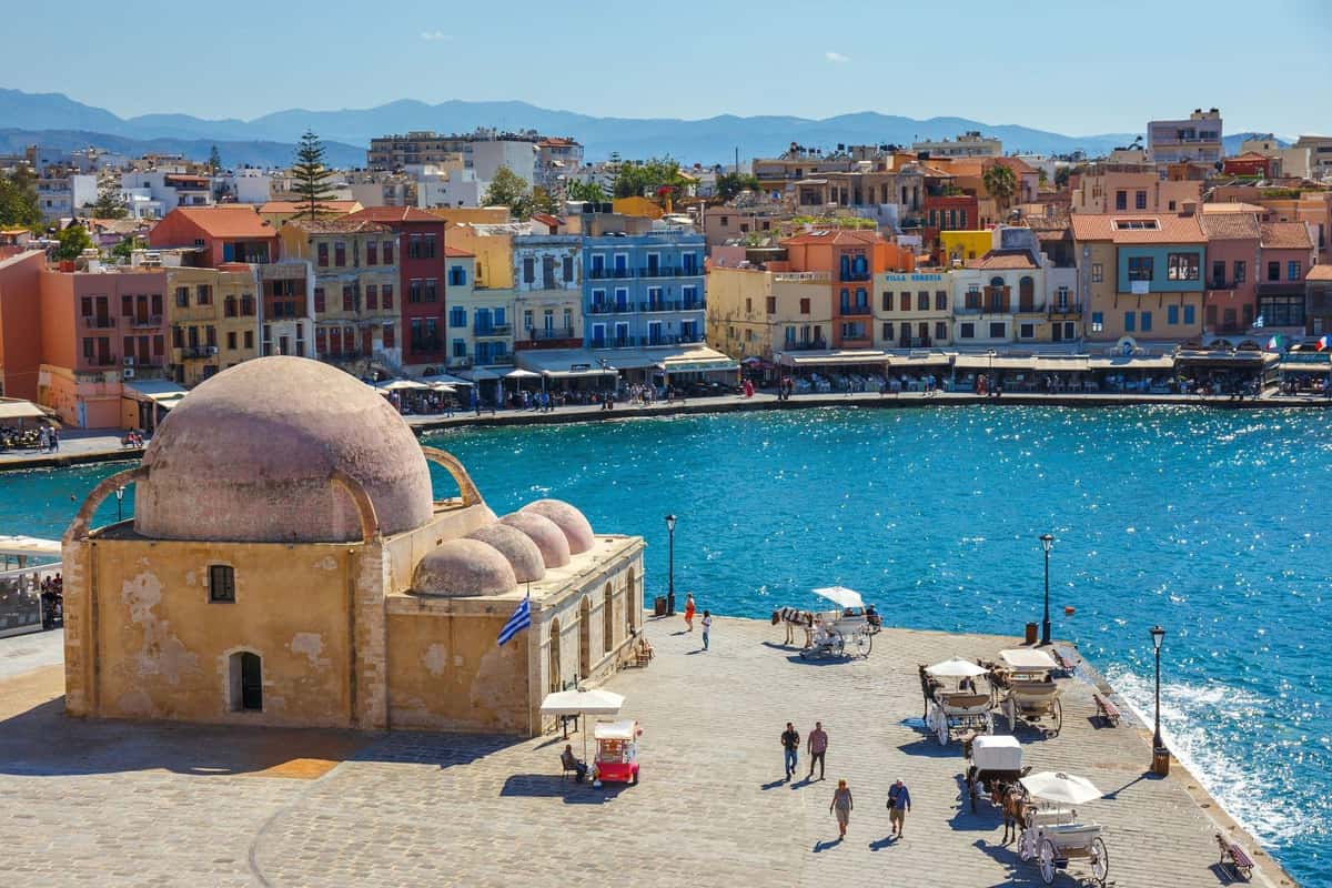 Chania food tour: the old harbour of Chania with a pale domed stone building on the quay, colourful multi-storey houses and cafe awnings along the waterfront, horse carriages and blue mountains beyond.