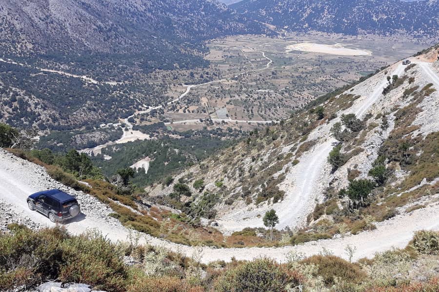 Chania jeep safari: a dark 4x4 drives along a white gravel track with sharp bends on a steep mountain slope above a wide valley of fields and trees, with a second vehicle on the track far above and bare grey mountains beyond.