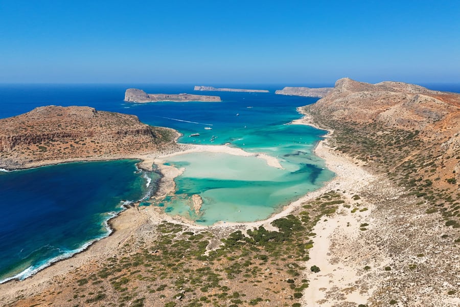 Chania tickets: an aerial view of a shallow turquoise lagoon with pale sandbars between a brown rocky headland on the left and a long sandy ridge on the right, deep blue sea beyond with small boats and low rocky islands on the horizon under a clear blue sky.