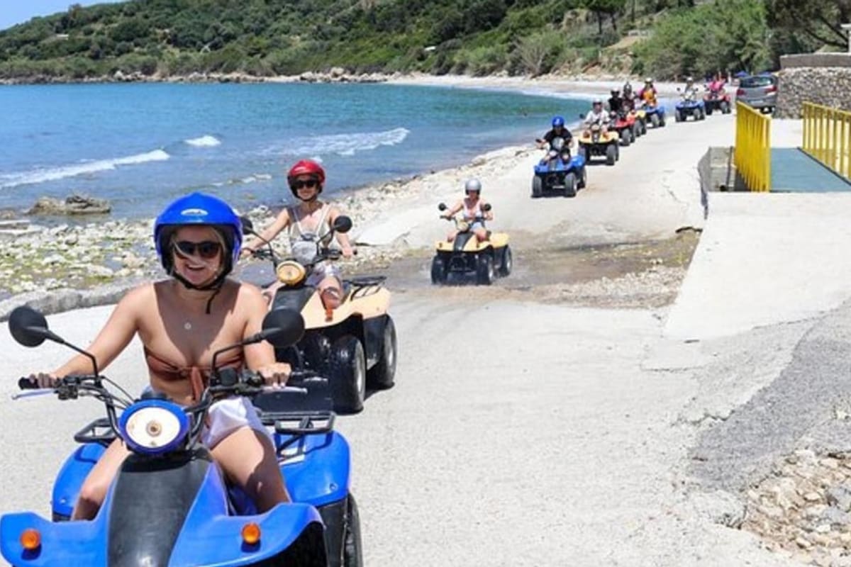 Corfu ATV tour: a line of quad bikes with helmeted riders rides along a pale coastal road beside clear blue sea, with a green wooded hillside across the bay under a bright sky.