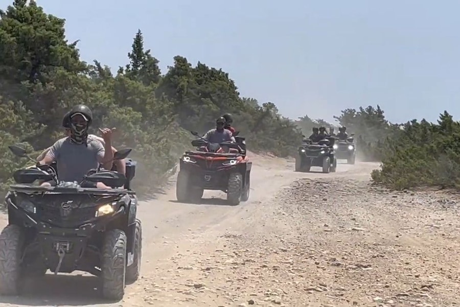 Corfu ATV tour: quad bikes with tour riders go in single file along a dusty gravel trail between green pine and scrub, with dust rising behind the riders under a hazy blue sky.
