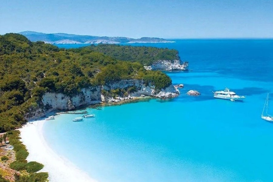 Corfu beach trip: a long white-sand beach curves around a bright turquoise bay below green wooded cliffs, with a white motor yacht and a sailboat anchored in the clear blue water.