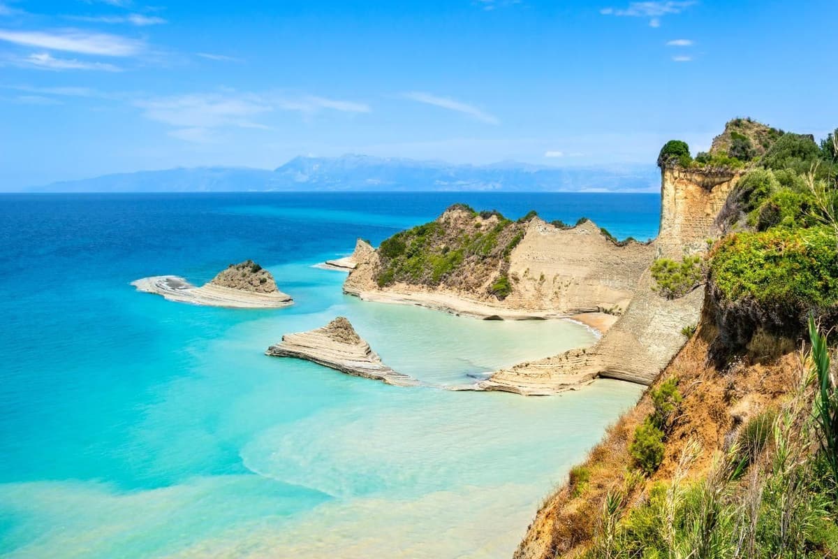 Corfu day trip: pale layered sandstone headlands and small rock stacks rise from a bright turquoise cove, with a green cliff top on the right and distant mountains across the blue sea under a clear sky.