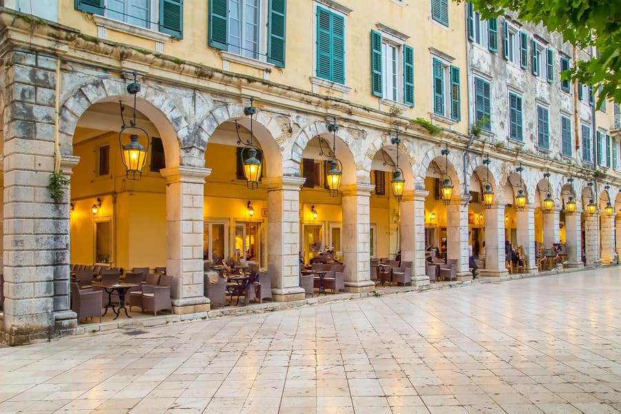 Corfu walking tour: a long row of wide stone arches with hanging iron lanterns runs along a pale yellow building with green shutters, with cafe chairs under the arcade and a smooth stone-paved square in front.
