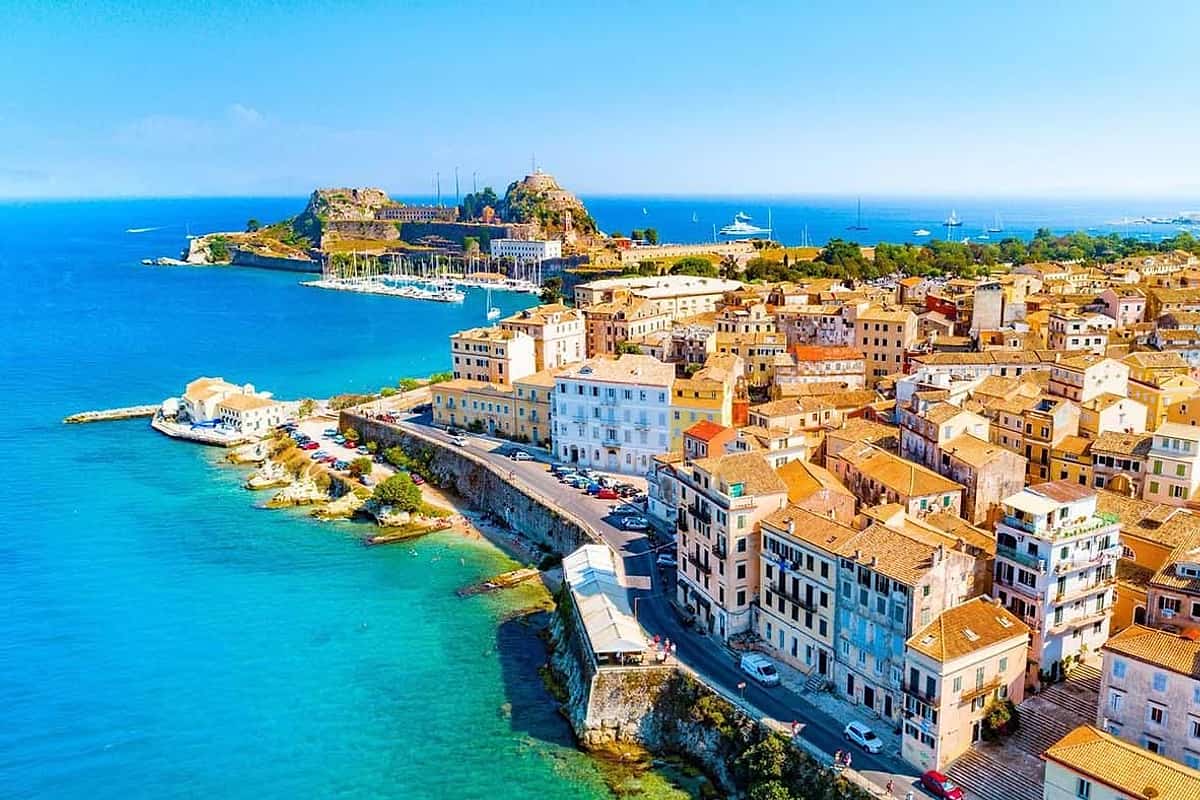 Corfu walking tour: an aerial view of a seafront old town of pale yellow, cream and orange buildings with tiled roofs, curving along a stone sea wall and a coastal road beside bright turquoise water, with a rocky fortress on a green headland, a marina of sailboats and a deep blue sea beyond under a clear sky.