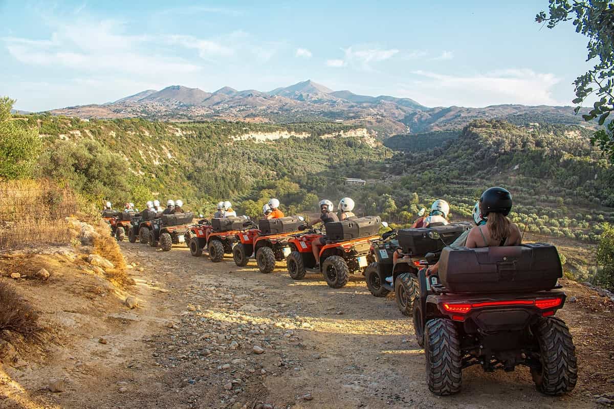Crete ATV tour: a line of orange and dark quad bikes with helmeted riders drives along a stony dirt track on a hillside above a green valley of olive groves, with a mountain range under a pale blue sky in warm evening light.