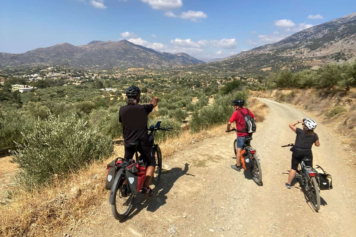 Crete bike tour: three e-bike riders with panniers, seen from behind, stop on a pale gravel track above a valley of olive groves with white houses and bare mountains under a blue sky with white clouds.