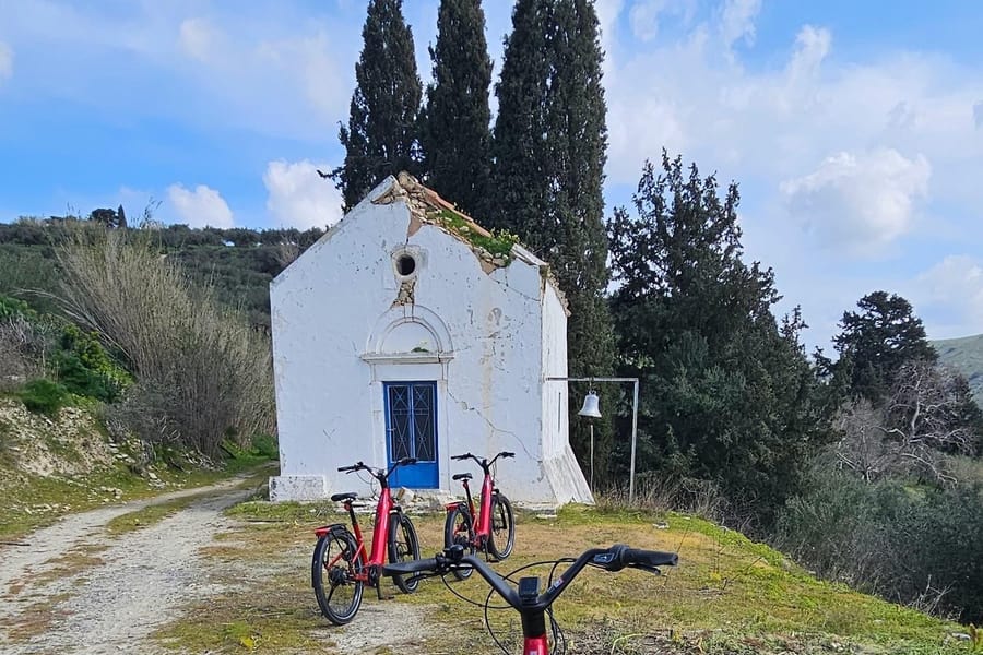 Crete bike tour: three red e-bikes parked on a grassy bank beside a gravel lane in front of a small whitewashed chapel with a blue door, a hanging bell and tall cypress trees under a blue sky.