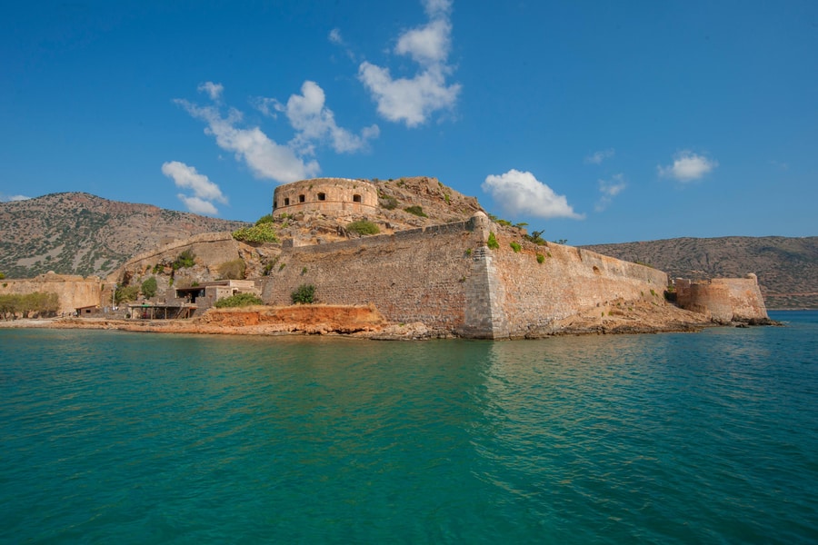 Crete boat trip: a thick-walled stone fortress with a round tower stands on a rocky islet above clear turquoise sea, under a blue sky with white clouds.