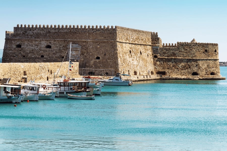 Crete castle tour: a large honey-coloured stone sea fortress with crenellated walls, small arched openings and a low corner tower stands at the edge of calm turquoise water, with small white fishing boats moored along the quay at the left under a pale blue sky.