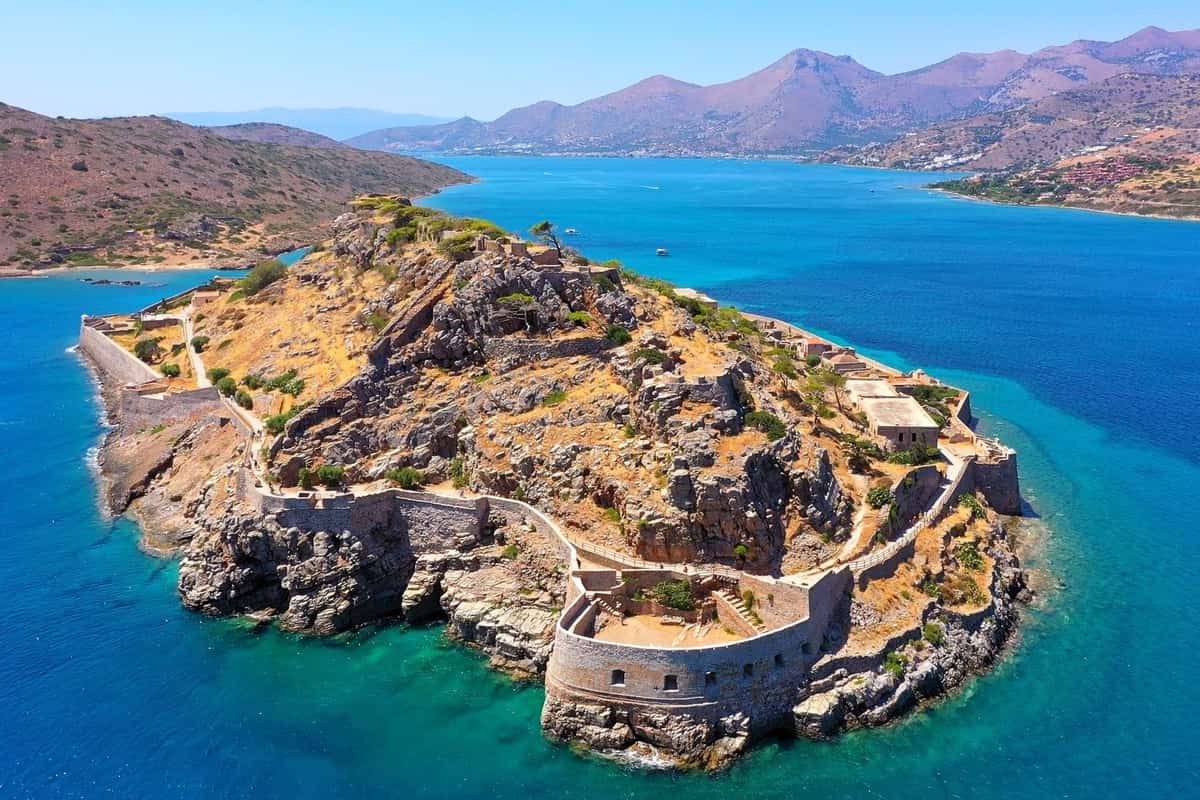 Crete castle tour: an aerial view of a stone fortress island with a curved bastion wall along its rocky sea edge, a ruined hilltop with terraced stone walls, deep blue and turquoise sea around it and brown mountains across the bay under a hazy blue sky.