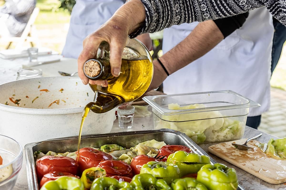 Crete cooking class: a hand tips a glass bottle of golden olive oil in a thin stream over a metal tray of red tomatoes and green peppers with cabbage leaves, beside a white bowl of leftover filling, a clear tub of sliced cabbage and a wooden board on a white worktable in daylight.
