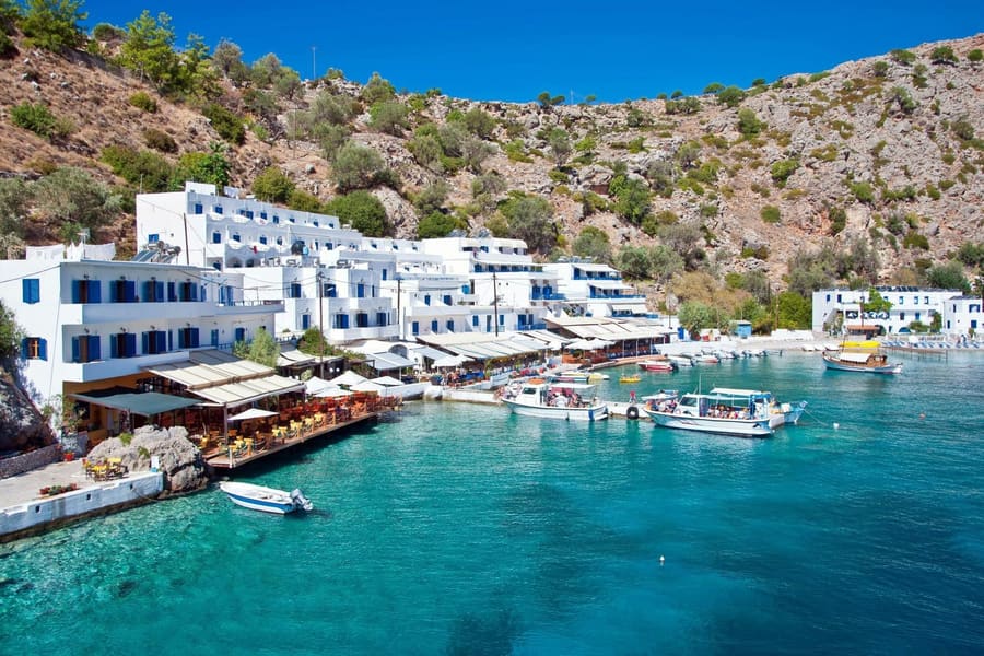 Crete day trip: white and blue buildings and a waterfront terrace with tables and umbrellas line a small bay of clear turquoise water, with several small boats moored at a quay below dry rocky hills and a blue sky.