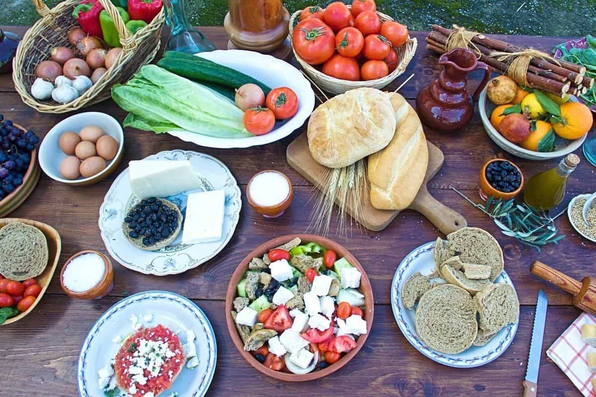 Crete food tour: a wooden table seen from above is laden with two loaves of bread on a board, baskets of red tomatoes and onions, a bowl of eggs, a salad with white feta cubes, plates of dark rusks and feta with black olives, a bottle of olive oil and bowls of grapes and oranges.