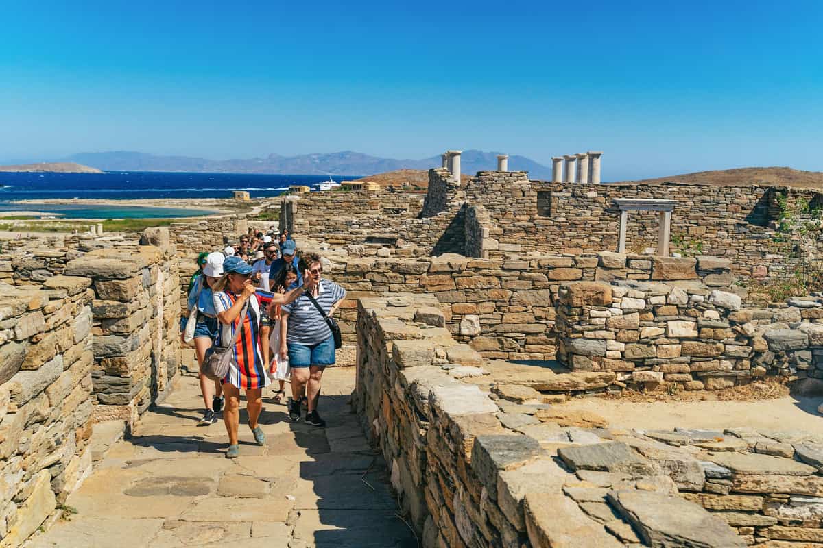 Delos tour from Mykonos: a small group of visitors walking through the ancient ruins of Delos with the blue sea behind