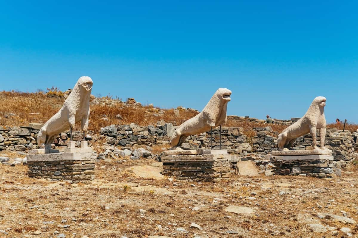 Delos transfer: the Terrace of the Lions on Delos, three archaic white marble lions in a row on stacked stone bases among low stone ruins and dry golden grass, under a clear blue sky.