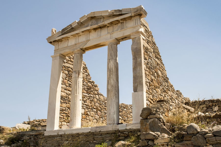 Delos transfer: a white marble gateway with a triangular pediment, two fluted columns and two square pillars stands in front of a tall stone wall, with a low stone base below and a clear blue sky above.