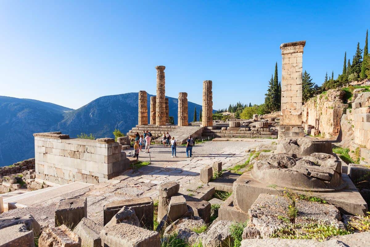 Delphi day trip from Athens visitors on the terrace of the Temple of Apollo under a blue sky with mountains behind