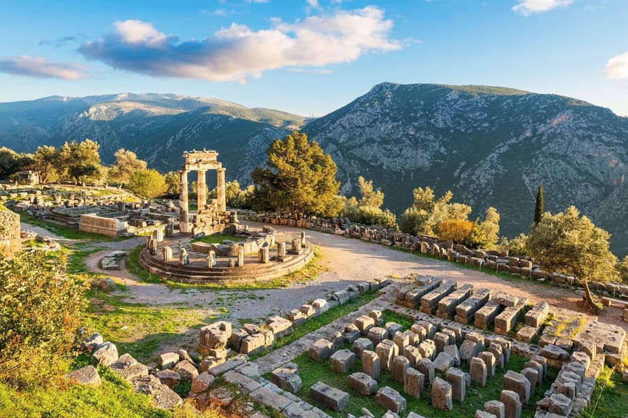 Delphi Meteora 2 day tour: the Tholos of Delphi, a round ruin with three standing columns on a stepped stone platform, stands in golden light among olive trees, with the stone foundation blocks of a temple in the foreground and mountains and a blue sky with clouds behind.
