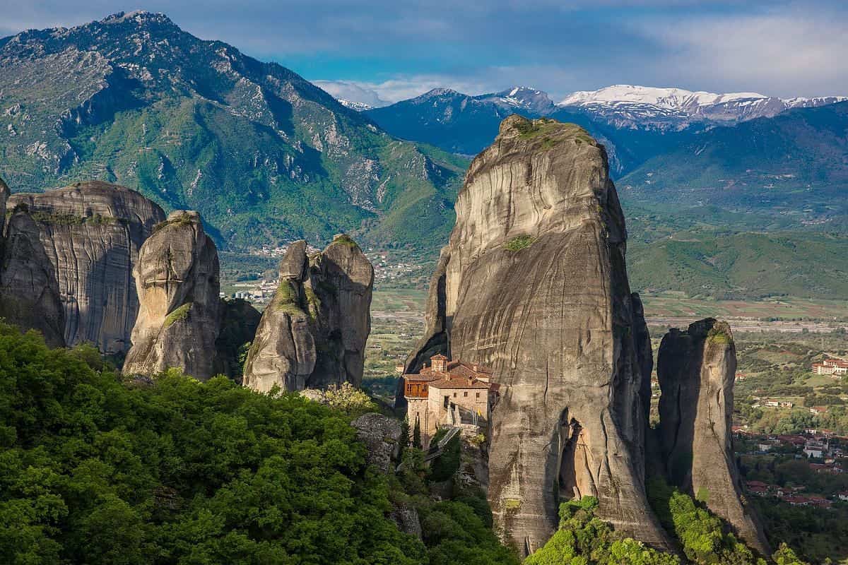 Delphi Meteora 2 day tour: a stone monastery with red-tile roofs stands on a rock ledge beside a huge sunlit rock pillar at Meteora, with more tall rock pillars, green forest, a valley with villages and snow-capped mountains behind it under a blue sky.