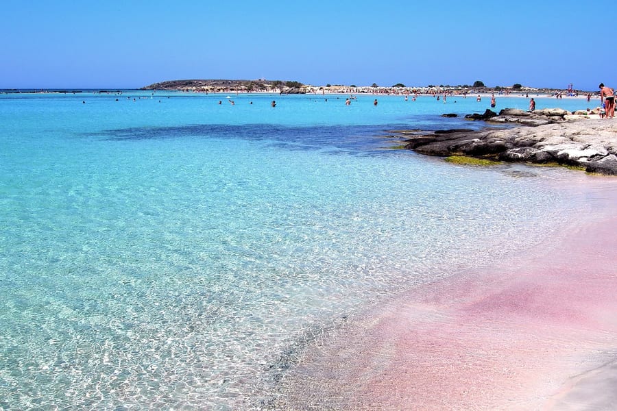 Pink sand and clear turquoise shallows at the water's edge on an Elafonisi day trip, with swimmers and the islet behind