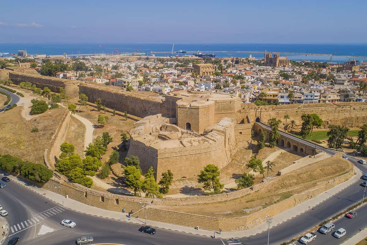 Famagusta ghost town tour: an aerial view of a huge sandstone bastion with thick walls and a stone arched bridge, ringed by green pines and a curving road, with white and orange-roofed houses, a harbour and deep blue sea behind.