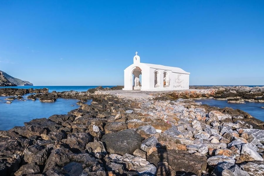 Georgioupolis land rover safari: the small white Agios Nikolaos chapel of Georgioupolis stands at the end of a rocky causeway between two pools of calm blue sea, under a clear blue sky, with a green headland on the left.